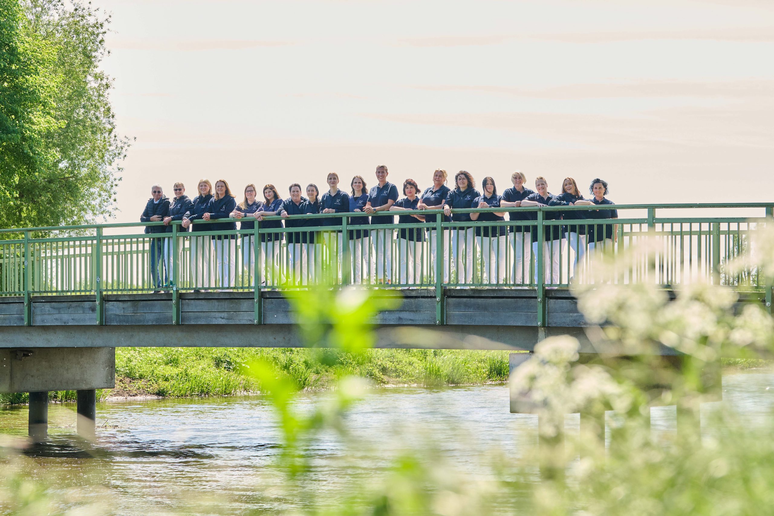 Eine Gruppe von Menschen steht Seite an Seite auf einer grünen Fußgängerbrücke über einen Fluss, mit Grün und Bäumen im Hintergrund.