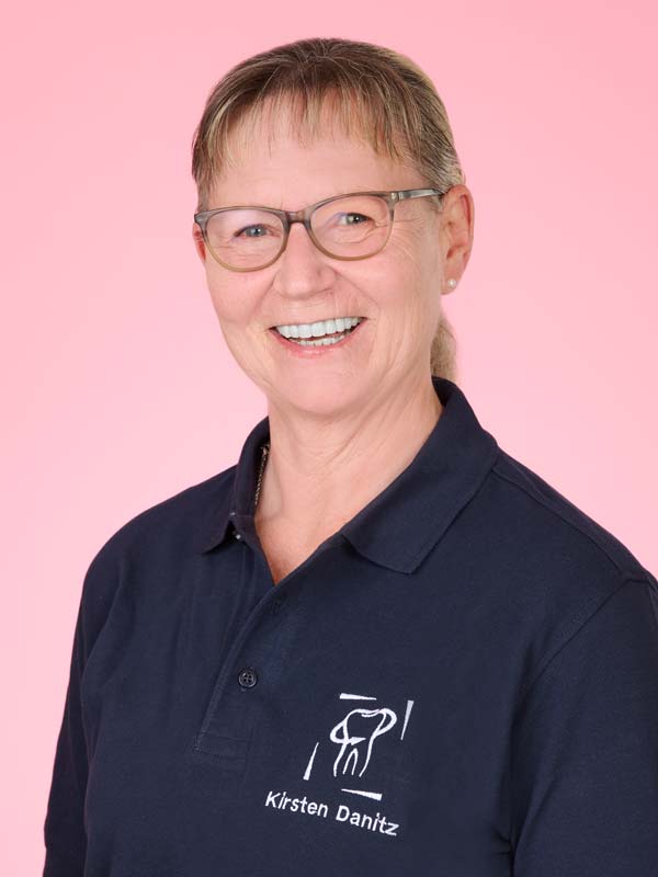 Eine lächelnde Frau mit Brille und einem marineblauen Poloshirt mit einem Zahnlogo und dem Namen "Kirsten Danitz" steht vor einem rosa Hintergrund.