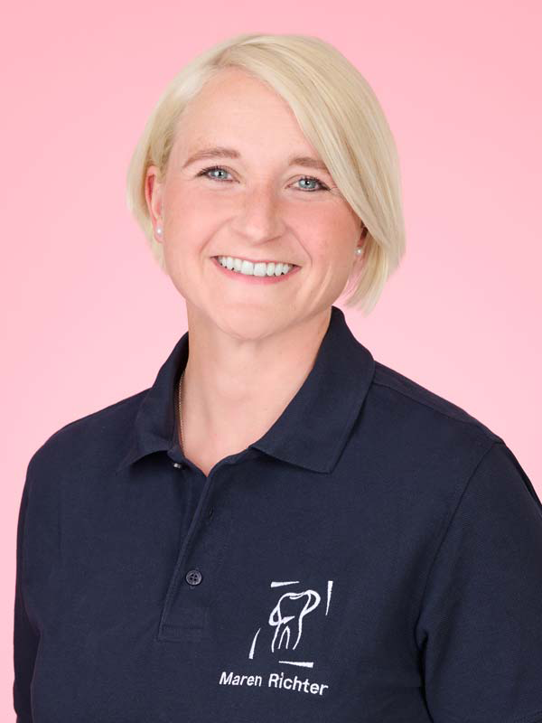 Eine lächelnde Person mit kurzen blonden Haaren, die ein marineblaues Kragenhemd mit einer Grafik und dem Namen "Maren Richter" trägt, steht vor einem rosa Hintergrund.