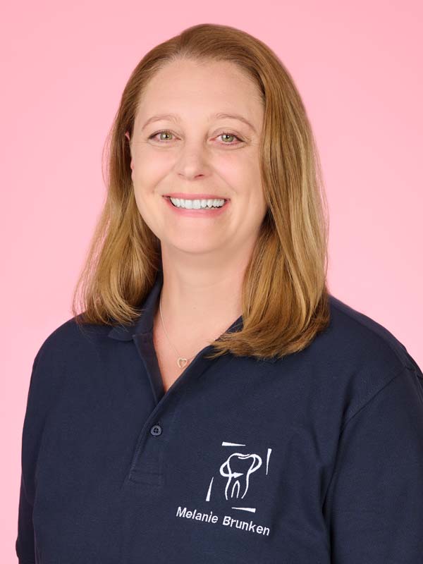 Eine Frau mit glattem, schulterlangem hellbraunem Haar lächelt in die Kamera. Sie trägt ein marineblaues Poloshirt mit einem Zahnlogo und der Aufschrift "Melanie Brunken".