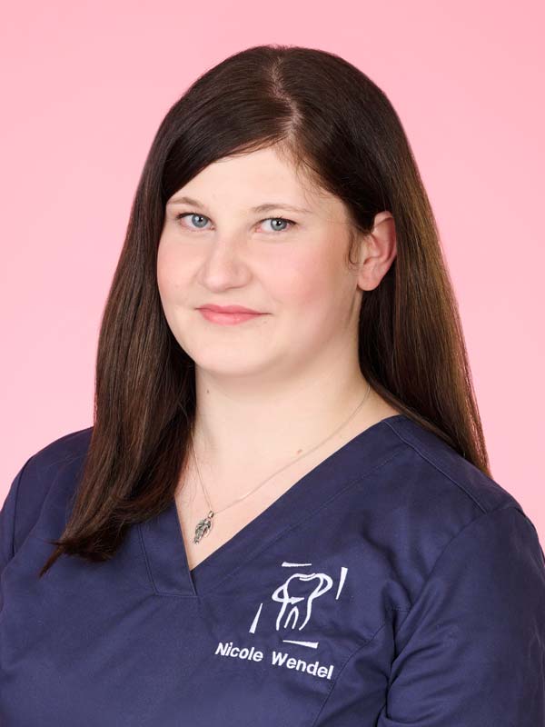 Frau mit glattem braunem Haar, die einen marineblauen Arztkittel trägt und vor einem rosa Hintergrund steht. Der Name "Nicole Wendel" ist auf ihr Oberteil gestickt.