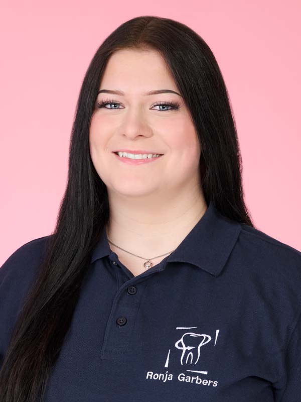 Eine Frau mit langen dunklen Haaren lächelt, sie trägt ein marineblaues Poloshirt mit einem Zahnlogo und dem aufgestickten Namen "Ronja Garbers" und posiert vor einem rosa Hintergrund.