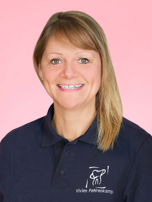 Lächelnde Frau vor einem rosafarbenen Hintergrund, die ein marineblaues Poloshirt mit einem Zahnlogo und dem aufgestickten Namen "Vivien Fehrenkamp" trägt.
