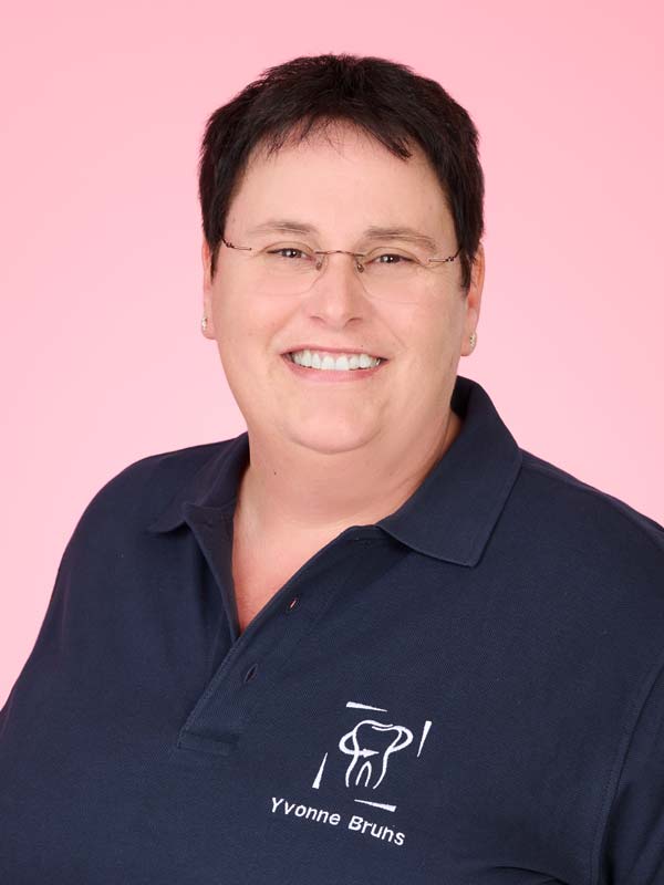 Eine Person mit kurzen dunklen Haaren und Brille lächelt, sie trägt ein marineblaues Poloshirt mit einem Dentallogo und dem Namen "Yvonne Bruhs" auf hellrosa Hintergrund.