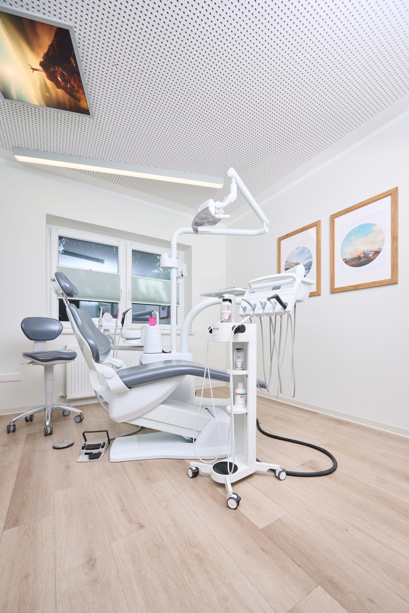 Eine moderne Zahnarztpraxis mit einem Zahnarztstuhl, zahnmedizinischen Geräten, hellem Holzfußboden und gerahmten Landschaftsfotos an der Wand.