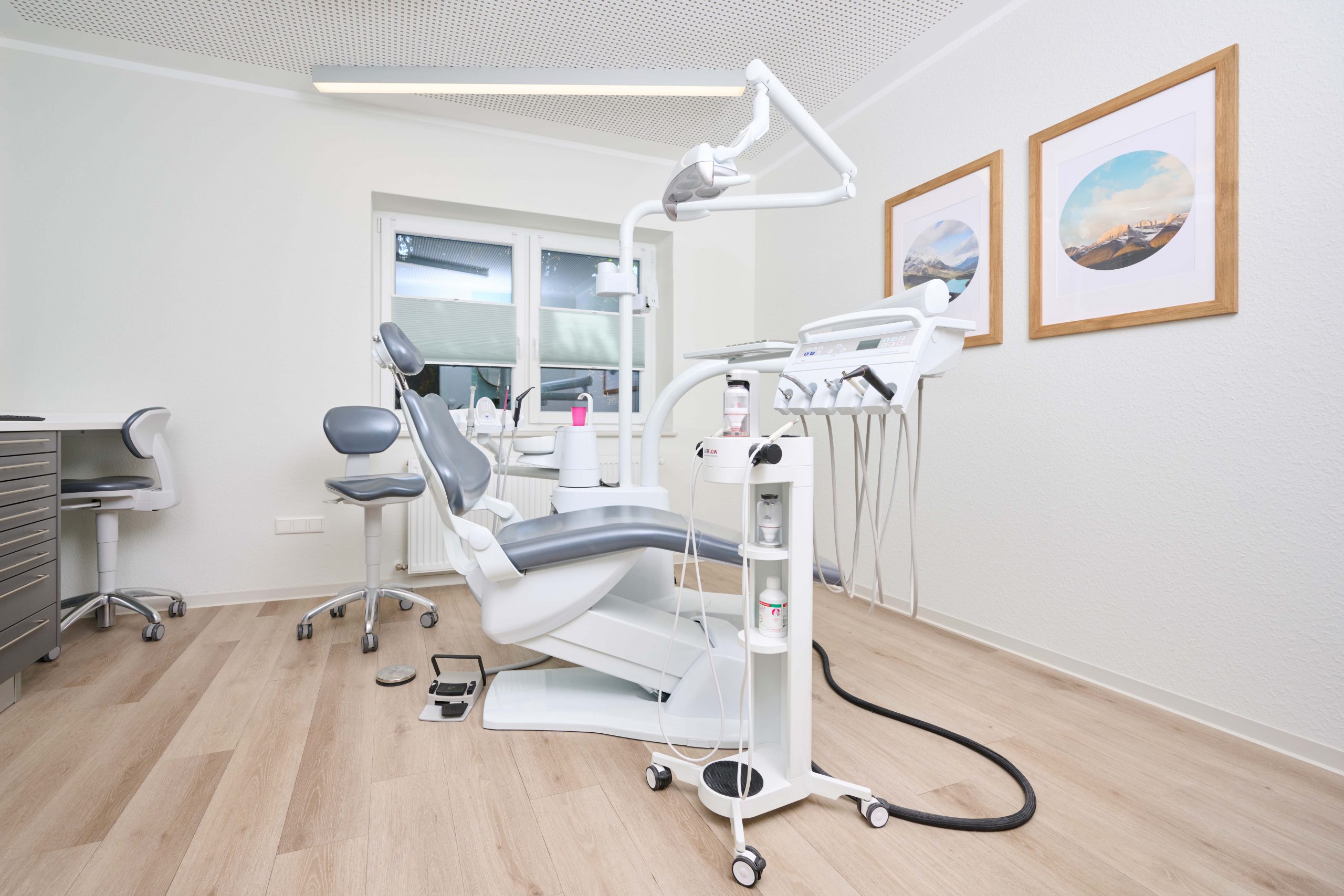 nicole-werthmann-zahnarzt_praxis_behandlungszimmer2 Eine moderne Zahnarztpraxis mit einem Zahnarztstuhl, Geräten und Werkzeugen in einem sauberen, hellen Raum mit Holzfußboden und gerahmten Bildern an der Wand.